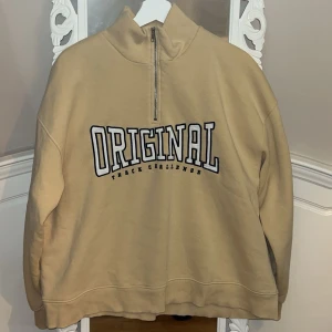 Beige tröja med dragkedja från Gina Tricot - Säljer en beige tröja från Gina Tricot med texten 'ORIGINAL' på framsidan. Tröjan har en halv dragkedja och långa ärmar, perfekt för en avslappnad stil. Den är gjord i ett mjukt material som känns skönt mot huden.