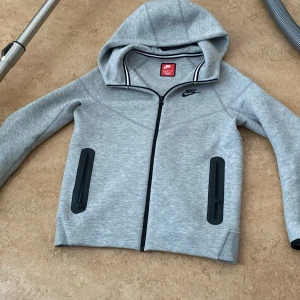 Grå huvtröja från Nike - Säljer en grå huvtröja från Nike med dragkedja och svarta detaljer. Tröjan har en justerbar huva och långa ärmar. Perfekt för en sportig look eller avslappnade dagar. 🏃‍♂️