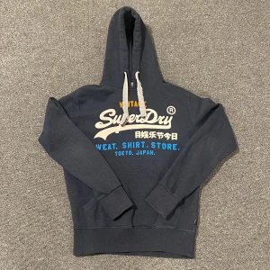 Mörkblå hoodie från SuperDry - Tja! Säljer en mörkblå hoodie från SuperDry med tryck i vitt, orange och blått på framsidan. Hoodien har en klassisk design. Perfekt för en avslappnad stil. Skick 9/10  Tveka inte med frågor!🙌