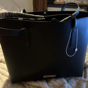 Svart axelväska från Anna Field - Snygg svart axelväska från Anna Field i syntetmaterial. Väskan har en stilren design med en praktisk dragkedja upptill. Perfekt för att bära med sig det nödvändigaste med stil. Väskan är helt ny som ni ser då plast sitter på, fick i studentpresent men inte min typ av stil🙌🏼🤩💗