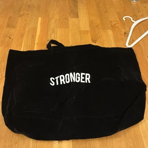 Svart shoppingväska från Stronger - Säljer en svart shoppingväska från Stronger med vit text på framsidan. Perfekt för att bära med sig allt man behöver under dagen. Väskan har en enkel design och är lätt att matcha med olika outfits.