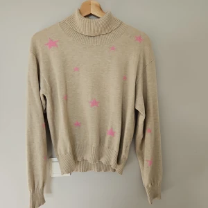 Beige polotröja med rosa stjärnor från Lindex - Säljer en beige polotröja från Lindex med rosa stjärnor. Tröjan har långa ärmar och ribbade muddar vid ärmslut och nederkant. Perfekt för kyligare dagar och ger en mysig känsla. 🌟