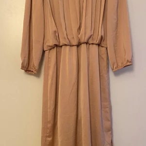 Beige klänning från Minimum Moves - Säljer en elegant beige klänning från Moves med långa ärmar och en draperad design framtill. Perfekt för en stilren look. Klänningen har en mjuk och flödande passform som ger en bekväm känsla.