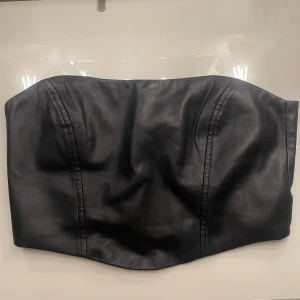Svart tubtopp från Nelly - Snygg svart tubtopp från Nelly i skinnimitation. Toppen har en dragkedja bak för enkel på- och avtagning. Perfekt för en stilren och edgy look. Är i nyskick använd 1 gång
