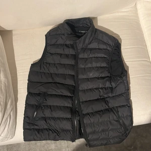 Svart dunväst från J.Lindeberg - Snygg svart dunväst från J.Lindeberg med dragkedja framtill. Perfekt för lager-på-lager under kyliga dagar. Västen har en stilren design med två fickor med dragkedja.