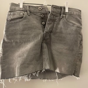 Grå jeanskjol från 157 - Snygg grå jeanskjol från 157 i storlek S. Kjolen har en rå kant och knappar framtill för en cool look. Perfekt för en avslappnad stil med fickor både fram och bak.