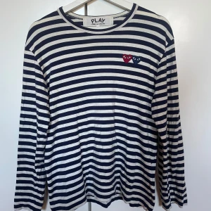 Randig tröja från Comme des Garçons - Säljer en randig tröja från Comme des Garçons med ikoniska hjärtbroderier i rött och blått på bröstet. Tröjan har långa ärmar och är i en klassisk marinblå och vit förgkombination. Storlek M.