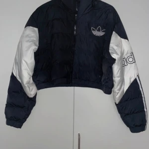Svart och vit pufferjacka från Adidas - Snygg svart och vit pufferjacka från Adidas med dragkedja och logga på bröstet. Jackan har långa ärmar och en croppad passform. Perfekt för en sportig look!