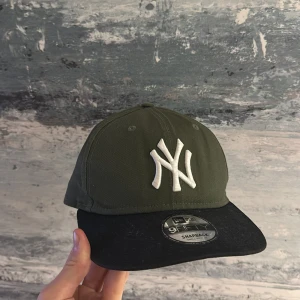 Grön och svart keps från New Era - Snygg grön keps med svart skärm från New Era. Kepsen har en broderad vit logga framtill och justerbar snapback-stängning baktill. Perfekt för en casual look.