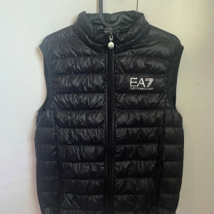 Svart dunväst från Emporio Armani - Snygg svart dunväst från Emporio Armani med dragkedja framtill. Västen har en quiltad design och en hög krage för extra värme. Perfekt för kyligare dagar när du vill hålla stilen.