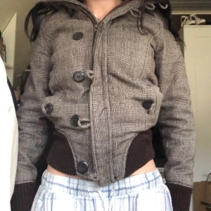 Brun rutig jacka med knappar y2k - Säljer en brun rutig jacka med stora knappar och dragkedja. Jackan har långa ärmar med ribbade muddar och en hög krage. Perfekt för höst och vinter!