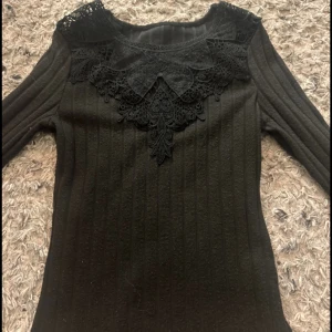 Svart ribbad tröja med spetsdetaljer - Snygg svart ribbad tröja med vackra spetsdetaljer vid halsen. Perfekt för en elegant look. Tröjan har långa ärmar och en åtsittande passform. Ett måste i garderoben för den som älskar stilrena plagg.