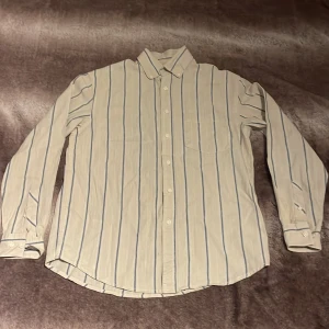 Grish Randig skjorta i linne/bomullsblandning från Cubus  - Säljer en stilren skjorta i linne och bomullsblandning från Cubus. Skjortan är randig i beige och blå med en avslappnadh passform. Storleken är s om man vill att den ska sitta lite större är man ute äftr en skjorta som sitter lite mer tajt och slimm då kan man köpa den om man är en storlek M Den har långa ärmar och knappar framtill. Perfekt för en avslappnad sommar grisch look