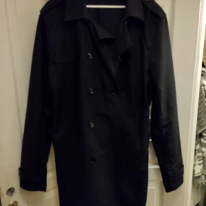 Svart trenchcoat - Snygg svart trenchcoat med knappar och bälte. Perfekt för höst och vår. Långärmad design med klassisk krage.