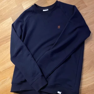 Mörkblå sweatshirt med broderad detalj - Säljer en stilren mörkblå sweatshirt med en liten broderad detalj i orange på bröstet. Tröjan har långa ärmar och en rund halsringning med ribbade muddar. Perfekt för en avslappnad look.