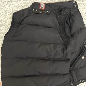 Svart dunväst från Parajumpers - Säljer en svart dunväst från Parajumpers med dragkedja och knappar. Västen har en quiltad design och en praktisk ficka med dragkedja på bröstet. Perfekt för kyligare dagar. En av de senare exklusiva modellerna, denna modell säljs ej kvar. Inga defekter och helt ny, max använd 3-4 ggr. Kvitto finns och kan mötas i Karlstad/Linköping, köpare står för frakt. Är strl L men passar för M.