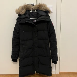 Svart dunjacka från Canada Goose - Säljer en svart dunjacka från Canada Goose med pälskantad huva. Jackan har långa ärmar och stängs med både dragkedja och knappar. Perfekt för kalla vinterdagar. 🖤