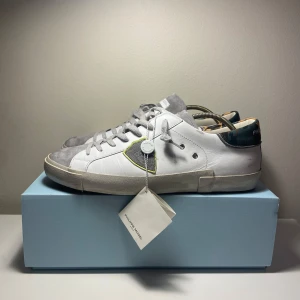 Vita sneakers från Philippe Model - Snygga vita sneakers från Philippe Model med grå detaljer och en unik camouflagemönstrad häl. Skorna har snörning och en diskret logga på sidan. Perfekta för en stilren look.