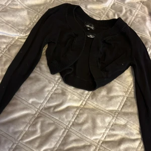 Svart bolerotopp från KappAhl - Säljer en stilren svart bolerotopp från KappAhl. Toppen har långa ärmar och en söt hjärtformad knapp framtill. Perfekt för att lägga till en extra touch till din outfit. Passar bra till både vardag och speciella tillfällen.