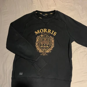 Svart sweatshirt från Morris - Snygg svart sweatshirt från Morris med ett stort tryck i guld på framsidan. Tröjan har långa ärmar och ribbade muddar vid ärmslut och nederkant. Perfekt för en avslappnad stil.