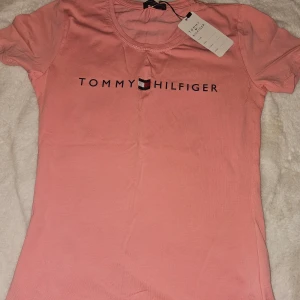 Rosa t-shirt från Tommy Hilfiger - Snygg rosa t-shirt från Tommy Hilfiger med logga på bröstet. Perfekt för en avslappnad stil. Kortärmad och i mjukt material för extra komfort.