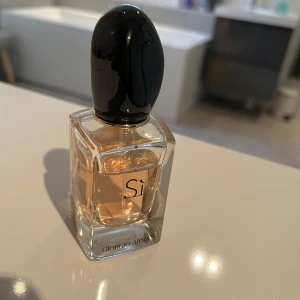 Si parfym från Giorgio Armani - Elegant parfymflaska från Giorgio Armani med doften Si. Flaskan är rektangulär och genomskinlig med en svart, oval kork. Den innehåller en ljus bärnstensfärgad vätska som ger en lyxig känsla. Perfekt för den som älskar sofistikerade och tidlösa dofter. 30ml