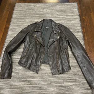 Brun skinnjacka från Trafaluc  - Snygg brun skinnjacka  med asymmetrisk dragkedja och stilrena detaljer. Jackan har en klassisk bikerstil med långa ärmar och flera fickor med dragkedjor. Perfekt för en cool look! Det står L på etiketten men den är en xxxs jacka 🫣