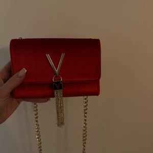Valentino väska - Snygg röd Valentino väska med guldfärgad kedja och dekorativ tofsdetalj. Perfekt för att ge en färgklick till din outfit.