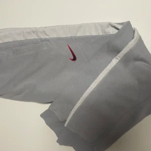 Grå mjukisbyxor från Nike - 10/10 skick.Nästan aldrig använda.Snygga grå mjukisbyxor från Nike med en vit rand längs sidorna och en röd Nike-logga. Byxorna har en bekväm passform och är perfekta för avslappnade dagar. De har elastiska muddar vid bensluten och en praktisk ficka. Size M.