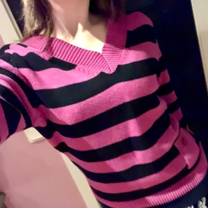 Fin V-ringad stickad tröja - Säljer en snygg rosa och svart randig tröja från cpm. Tröjan har en v-ringad hals och en liten ficka med knappdetalj på framsidan. Perfekt för en färgglad look!