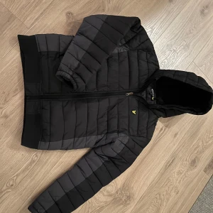 Svart pufferjacka från Lyle & Scott - Säljer en svart pufferjacka från Lyle & Scott med dragkedja och en gul logotyp på bröstet. Jackan har långa ärmar och är perfekt för kyligare väder. Den är designad i Storbritannien och passar ungdomar i åldern 14-15 år.