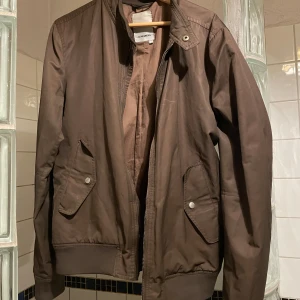 Brun jacka från J.Lindeberg - Snygg brun jacka från J.Lindeberg med dragkedja och knappar. Perfekt för höst och vår. Jackan har långa ärmar och ribbade muddar vid ärmslut och nederkant. Två fickor med knappstängning framtill.