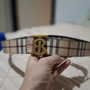 Rutig bälte från Burberry - Snyggt bälte från Burberry med klassiskt rutigt mönster i beige, svart och röd. Bältet har ett stort guldfärgat spänne med Burberrys ikoniska logotyp. Perfekt för att ge en lyxig touch till din outfit.