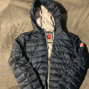 Blå pufferjacka från Colmar - Säljer en snygg blå pufferjacka från Colmar. Jackan har en dragkedja framtill och en praktisk huva. Perfekt för kyligare dagar med sin quiltade design som håller värmen. Den har en stilren look med Colmar-logga på ärmen.