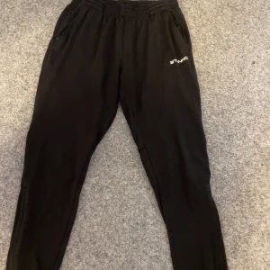 Svarta träningsbyxor från Stanno - Säljer ett par svarta träningsbyxor från Stanno. Byxorna har en elastisk midja och är tillverkade i ett syntetmaterial som är perfekt för träning. De har en diskret logga på ena benet och en bekväm passform.