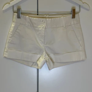 Vita shorts med knappar - Snygga vita shorts med uppvikta ben och två knappar framtill. Perfekta för varma sommardagar. De har en klassisk design med fickor på sidorna.