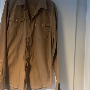 Beige långärmad skjorta med knappar - Snygg beige långärmad skjorta från Levi's med klassisk krage och knappar framtill. Skjortan har två bröstfickor med knappstängning och en diskret logga på ena fickan. Perfekt för en stilren och avslappnad look.Inte använd.