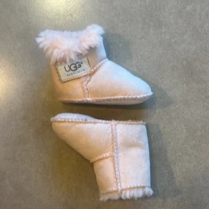Rosa babyboots från UGG - Söta rosa babyboots från UGG med mjukt foder och klassisk design. Perfekta för att hålla små fötter varma och bekväma. Tillverkade i mocka med fluffig pälsdetalj runt öppningen. I storlek S.