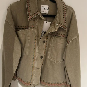 Beige jacka från Zara - Säljer en beige jacka från Zara med broderade detaljer i rosa och svart. Jackan har en croppad design med knappar framtill och fransar vid nederkanten. Perfekt för att ge din outfit en unik touch!