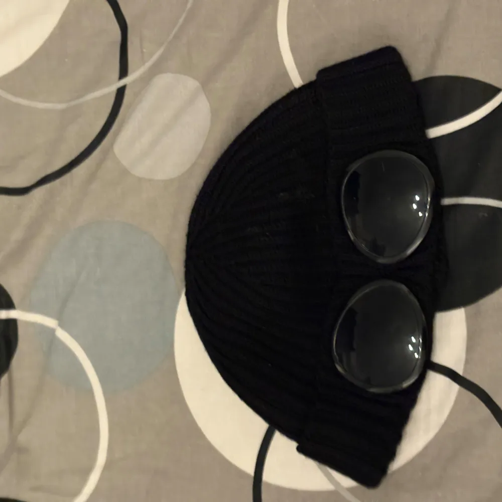 Snygg svart ribbad mössa från C.P. Company med unika glasögondetaljer framtill. Perfekt för att ge din outfit en cool touch. Tillverkad i ett mjukt och bekvämt material.. Asusteet.