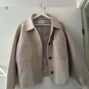 Beige ulljacka från NA-KD - Säljes pga kommer ej till användning. från NA-KD med stora fickor framtill och knappar. Jackan har en klassisk krage och en rak passform som ger en modern look. 