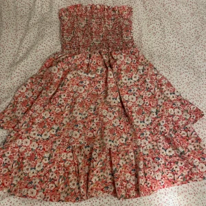 Blommig axelbandslös klänning - Charmig axelbandslös klänning med ett färgglatt blommönster i rosa och vita nyanser. Den har en smockad överdel som ger en bekväm passform och en luftig kjol som faller fint. Perfekt för varma dagar och somriga tillfällen.