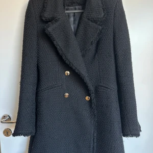 Svart kappa med guldknappar ZARA - Elegant svart kappa från ZARA med dubbelknäppning och fransiga detaljer vid ärmslut och krage. Perfekt för kyligare dagar med sin långa design och klassiska look. Storlek S