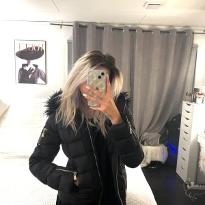 Svart dunjacka med pälskrage - Snygg svart dunjacka med fluffig pälskrage och dragkedjor på ärmarna. Jackan har en figurnära passform och är perfekt för kyliga dagar. Den har även en dekorativ patch på ärmen. Perfekt för att hålla stilen i vinter! 🖤