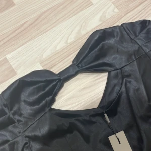 Svart klänning med rosett - Säljer en elegant svart klänning med en stor rosett vid halsen. Klänningen har en öppen rygg och är tillverkad i ett glansigt material som ger en lyxig känsla. Perfekt för festliga tillfällen!