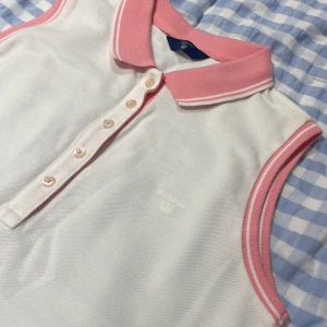 Vit ärmlös pikétopp från GANT - Säljer en snygg vit ärmlös pikétopp från GANT med rosa krage och kantdetaljer. Toppen har knappar framtill och är perfekt för en avslappnad men stilren look. Passar bra till både vardag och sommaraktiviteter.
