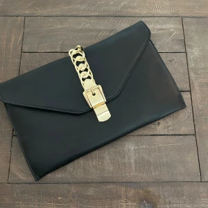 Svart kuvertväska med gulddetaljer - Snygg svart kuvertväska i syntetmaterial med en elegant guldkedja som detalj. Väskan har en praktisk dragkedja inuti för extra säkerhet. Perfekt för festliga tillfällen eller en utekväll när du vill ha något stilrent och enkelt. 