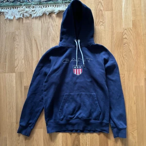Mörkblå hoodie från GANT - Säljer en snygg och bekväm mörkblå hoodie från GANT. Den har en klassisk design med en stor ficka fram och justerbar huva med vita snören. På bröstet finns GANT-loggan.