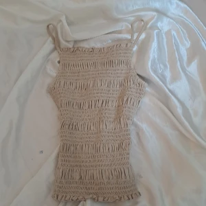 Beige smockad topp från H&M - Säljer en superfin beige smockad topp från H&M. Den är ärmlös med smala axelband och har en tight passform som är perfekt för sommaren. Materialet är mjukt och stretchigt, vilket gör den bekväm att bära hela dagen.