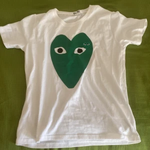 Vit t-shirt med grönt hjärta från Comme des Garçons - Säljer en snygg vit t-shirt från Comme des Garçons med ett stort grönt hjärta på framsidan. T-shirten är i bomull och har en normal passform. Perfekt för en avslappnad stil!